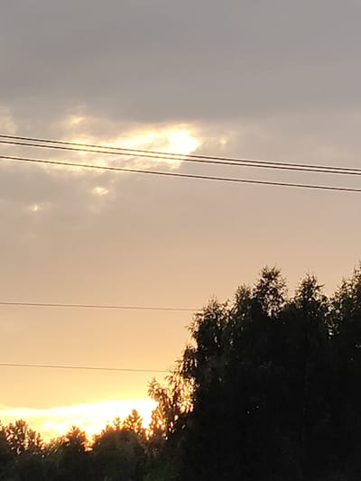 This media file depicts a natural landscape scene, likely a rural area given the context of Skurygino, Russia. The primary subjects are the sky, clouds, trees, and overhead power lines. The time of day is clearly indicated as sunset or sunrise, with the sky exhibiting warm hues of orange and yellow, transitioning to a dusky gray towards the top. The sun's rays are piercing through gaps in the clouds, creating a luminous effect.
The trees form a silhouetted border along the bottom and right edges of the frame, suggesting a forest or a line of trees bordering a field. Their dense foliage is dark, contrasting with the bright sky. Three parallel power lines traverse the upper portion of the image horizontally, indicating human infrastructure within the natural setting.
The weather appears to be overcast with some clearing, allowing the sunlight to break through. There are no people or specific objects of interest, with the focus being on the atmospheric conditions and the interplay of light and shadow. The overall mood evoked is one of tranquility and natural beauty, characteristic of a serene evening or morning in the countryside. No text is visible in the image.