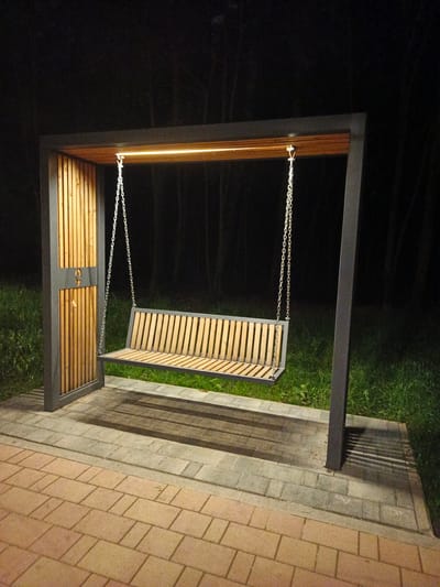 In Sof'ino, Russia, a modern, illuminated park bench swing is the central subject of this nighttime photograph. The swing, constructed from metal with a wooden slatted seat and backrest, hangs from a dark grey metal frame. Integrated warm LED lighting illuminates the top of the frame. The frame is supported by two vertical posts; one is adorned with vertical wooden slats and a small, circular emblem. The swing is situated on a paved area composed of interlocking brick pavers in shades of grey and orange, bordering a grassy area. In the background, a dark, out-of-focus forest serves as the environmental backdrop. The lighting casts distinct shadows of the swing's chains and frame onto the paved surface.