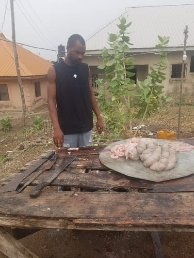 A man in a black sleeveless shirt and blue jeans stands in front of a wooden table with two large knives and a plate of raw meat on it. He is looking down at the meat. Behind him is a small house with a corrugated metal roof. There is a green bush to the right of the man. The scene is in Pati Ridge, Nigeria, at the coordinates latitude: 7.81, longitude: 6.704.  The table is made of weathered wood planks. There is a small amount of blood on the table and on the floor. The meat is a light pink color and appears to be innards.  The man is not smiling and seems to be concentrating on the task at hand.