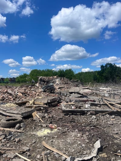 The image shows the aftermath of a destroyed building in Terny, Ukraine. The building appears to be made of concrete blocks and has been reduced to rubble. The debris is scattered across the ground, mixed with broken wood and other materials. There are large, white cumulus clouds in the bright blue sky. The site is covered in debris and overgrown with vegetation. There are no people in the image. The coordinates indicate that this is a location in a forested area. The scene appears to be a result of some type of significant destruction, potentially from a bombing or other attack.