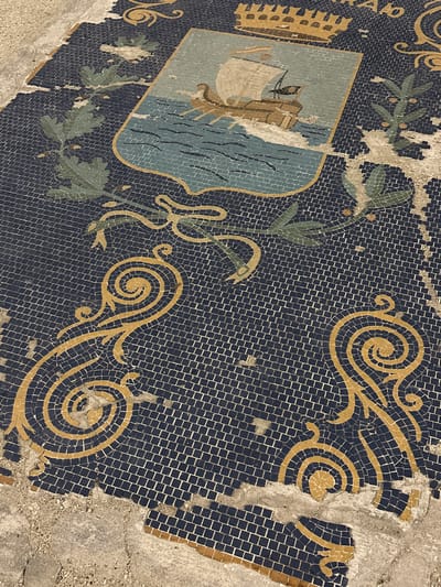 This media file depicts a detailed and partially damaged mosaic, likely a coat of arms or emblem, set into a dark blue tiled background. The main subject is a shield-shaped motif on the left, rendered in shades of light blue, beige, and brown. Within the shield, a stylized ship with a sail floats on blue waves, symbolizing maritime activity. Above the shield, a golden crown or fortified structure, possibly a city wall, is visible.

Surrounding the central emblem are elaborate decorative patterns. Golden yellow tesserae form intricate swirls and curvilinear designs that extend across the dark blue background. Interspersed among these golden elements are green foliage patterns, resembling branches with leaves.

The mosaic shows significant wear and damage, with numerous tesserae missing in various sections, particularly along the edges and within the decorative elements. The exposed areas reveal a rough, greyish concrete or mortar substrate underneath. The perspective is an angled overhead shot, emphasizing the texture and three-dimensionality of the mosaic tiles.

No specific activity or event is taking place; the image is a static portrayal of an artistic installation. The overall impression is one of historical artistry suffering from the passage of time. The location context of Portoferraio, Italy, suggests this mosaic could represent the city's heraldry, given its historical significance as a port. Faint, partial letters, possibly "Ab" or similar, are visible in the top left corner, though largely indistinct due to damage.