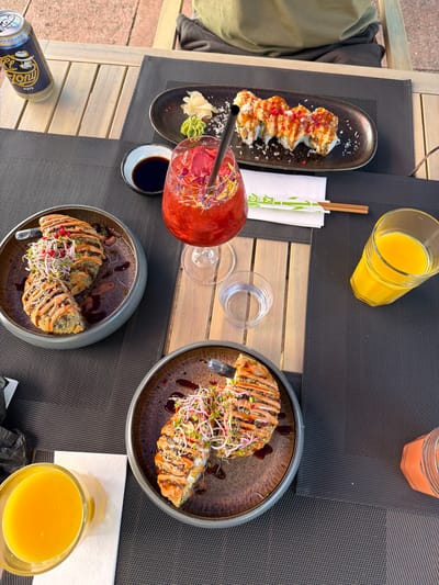 The image depicts an outdoor dining table set with various food and drink items on a sunny day. The table is constructed from light-toned wooden slats and covered with dark gray woven placemats.
In the upper center, an oval-shaped dark plate holds eight pieces of sushi rolls, each topped with a light-colored sauce, red granular sprinkles, and white flakes. To the left of the sushi, a small black plate presents a mound of pickled ginger, a portion of green wasabi paste, and a white grated condiment. A small white bowl containing dark soy sauce is positioned nearby.
A tall glass, centrally located, contains a vibrant red cocktail adorned with red flower petals and small blue/purple floral elements, with a black straw inserted. An "EL TONi MATE" beverage can, blue and gold with a graphic of a man in a hat, is situated in the upper left corner.
Two individual ceramic bowls, colored dark gray with a mottled interior, each contain large, oblong, deep-fried or tempura-battered rolls. These rolls are sliced and drizzled with a light tan sauce and a darker, possibly balsamic-based, glaze. Both portions are garnished extensively with green and purple sprouts and scattered red peppercorns. One bowl is prominent in the mid-left, and the other is in the lower right.
Additional beverages include two clear glasses of orange juice, one in the mid-right and another in the lower left. A small clear glass of water is also on the table. A white napkin, folded and featuring green Japanese characters, holds a pair of wooden chopsticks. A partial view of an orange-colored cylindrical object, possibly a cup or container, is visible in the lower right.
A person wearing a khaki or olive-green shirt is partially visible sitting in the background. The environment suggests an outdoor restaurant or cafe terrace, with a light-colored building and reddish-brown paved ground visible in the distant background.
Based on the distinct culinary presentation, particularly the style of the large fried rolls and sushi, combined with the presence of the Swiss-brand "EL TONi MATE" drink, the scene is highly consistent with the offerings and typical outdoor dining setting of the "Mitte" restaurant, located in Basel, Switzerland.