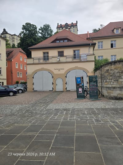 This is a vertical, eye-level photograph taken in Pirna, Germany, on June 23, 2026, at 10:12 AM. The scene depicts the exterior of a historical building, identified by the text "Feuerlöschgerätehaus 1914" as a fire equipment house built in 1914. The building has a light yellow facade with large, arched double doors painted white. Above the doors, a sign reads "Uniwerk" and "www.uniwerk.de" indicating a connection to an organization or event called Uniwerk. A bicycle is parked next to a chalkboard sign which lists event details and website information. The building is situated on a cobblestone street. In the background, other buildings with red-tiled roofs and traditional architecture are visible, as are trees and a stone wall. The sky appears overcast, suggesting a cloudy day. The overall impression is one of historical architecture and a public space possibly hosting an exhibition or event.