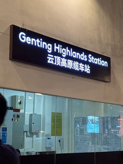 The image depicts a sign for "Genting Highlands Station" with its Chinese translation. The sign is a black rectangle with white illuminated text. It's mounted on a wall. Adjacent to the sign is a glass panel displaying a white board with writing and a blue square, possibly a projection or screen. Below the whiteboard, there are electrical boxes and panels. A person's head is partially visible in the lower center of the frame. The environment suggests an indoor setting. The location is Genting Highlands, Malaysia, as indicated by the sign.