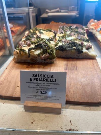The image captures a close-up view of two rectangular slices of "Salsiccia e Friarielli" pizza displayed on a light-colored wooden cutting board, likely within a glass food display case in a bakery, pizzeria, or deli in Dorno, Italy. The pizza slices feature a thick, airy crust, topped with mozzarella fiordilatte cheese, crumbled sausage, and cooked rapini (broccoli rabe).
To the right of the pizza, a white price and description tag is prominently displayed, held upright by a small wooden stand. The tag provides details in both Italian and English. The main title reads "SALSICCIA E FRIARIELLI." Below this, the Italian description states "Pizza alla pala con mozzarella fiordilatte, salsiccia e friarielli," followed by the English translation: "Pizza alla pala with fiordilatte mozzarella, sausage and rapini." The price for this pizza is clearly marked as "€ 8,20."
Further text on the tag indicates, "Elenco completo ingredienti, allergeni e caratteristiche su QR code dedicato" (List of ingredients, allergens and characteristics on dedicated QR code) and "Prodotto con base congelata all'origine" (Product with base frozen at source). The setting appears to be indoors with artificial lighting, showcasing the prepared food for sale. Part of another pizza or baked good is visible in the upper left background, suggesting a variety of items are available.