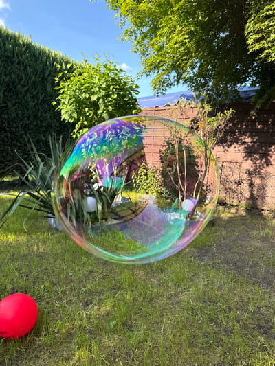 The image depicts a large, iridescent soap bubble floating in an outdoor setting. The bubble reflects the surrounding environment, including green trees, a wooden fence, and a blue sky. A red ball sits on the grass near the bottom right corner of the frame. The location is Lauchhammer, Germany, based on the presence of a building and other environmental variables that are known to be located in that city.