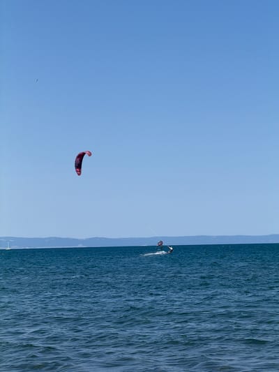 The image depicts a bright, clear day over the sea, likely the Black Sea coast near Atiya, Bulgaria. The sky is a vivid, cloudless blue. The sea itself is a deep blue, with small, gentle waves creating a textured surface.
In the middle ground, two individuals are kiteboarding. The kiteboarder on the left is larger in the frame and more prominent. They are utilizing a kite with a distinctive red and purple checkered pattern, which is curved and airborne, pulling the rider across the water. The rider is leaning back, engaged in the sport.
Further to the right and slightly more distant, another kiteboarder is visible. This rider is smaller in the frame and appears to be actively riding on the water, with their kite, a red one, also in the air.
In the far background, a low, hazy line of land with rolling hills is visible, representing the coastline. A faint white sail of a boat can be seen on the far left, hinting at other maritime activity. A single bird is present in the upper left quadrant of the sky. The overall scene is one of active recreation on a sunny day at the coast.