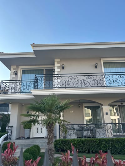 The image shows a modern building with multiple stories, featuring balconies with decorative wrought iron railings and large windows. The facade is a light gray color with white trim. A palm tree and manicured hedges are visible in the foreground, suggesting a warm climate. The sky is clear and blue. The scene appears to be a residential or possibly commercial property in Chernomorets, Bulgaria, given the architectural style and landscaping often found in coastal areas of the country. There are no people or specific actions depicted in the image. The overall impression is one of a well-maintained, upscale property.