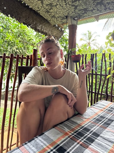 An adult female is depicted seated at a table on an outdoor veranda in Nilwella, Sri Lanka. She has blonde hair, with sunglasses resting atop her head, and is wearing a light-colored t-shirt. Her left forearm displays a circular mandala-style tattoo, and her right forearm bears Roman numerals "XIX.X.IX". She wears large, amber-colored, butterfly-shaped earrings. She is holding a lit cigarette between the index and middle fingers of her right hand, from which smoke is visibly rising. Her eyes are partially closed, and her mouth is slightly open, consistent with exhaling smoke. Her left leg is bent, with her left forearm resting on her knee. The table is covered with a plaid tablecloth featuring black, white, and orange squares. The background consists of lush green tropical foliage, including trees and a palm visible against a bright, overcast sky. A rustic fence made of vertical wooden poles is present behind her. Above, a patterned fabric ceiling is visible, along with a woven mat ceiling section and a support column wrapped in reddish material from which a potted plant hangs.