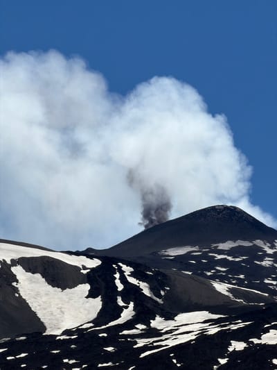The image depicts Mount Etna, a volcano located near Linguaglossa, Italy. The upper portion of the image shows a clear blue sky with a large plume of white and gray smoke emanating from the peak of the volcano. The volcanic cone is dark and barren, with patches of snow scattered across its slopes. The foreground is dominated by dark volcanic rock, also interspersed with snow. The scene captures a moment of volcanic activity.