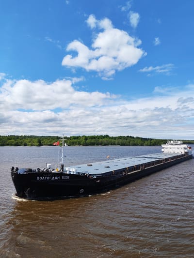 A barge, the "Волго-Дон 5051" ("Volgo-Don 5051"), travels on a river. The barge is dark in color with a lighter-colored, flat top and a small white structure on the rear. The river is brown and reflective, showing ripples and waves. The river bank in the background is covered in green trees. The sky overhead is blue with scattered white clouds. The location is near Popovo, Russia, based on the barge's name, which refers to the Volga-Don Canal, a Russian waterway.