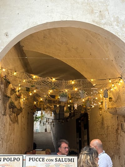 The image captures an atmospheric, arched stone passageway in Polignano a Mare, Italy, adorned with nautical-themed decorations. The passage, characterized by its rough, textured stone walls and rounded ceiling, is illuminated by numerous warm string lights woven into fishing nets that hang overhead, creating a canopy effect. These nets are decorated with small wooden fish, anchors, steering wheels, and miniature woven baskets, enhancing the coastal ambiance.
Several people are visible within the passage. In the mid-ground on the right, a man with a bald head stands next to a woman with long, wavy blonde hair. Closer to the foreground, a man with glasses peers towards the camera from behind a menu board. Other indistinct figures can be seen further down the narrow passage. The artificial lighting suggests either evening or a dimly lit indoor/covered space, contributing to a cozy and inviting atmosphere.
Visible text on menu boards on the right-hand side indicates food offerings. One sign reads "N POLPO A MONACHILE Polpo." Another prominent sign advertises "PUCCE CON SALUMI" (Pucce with Cured Meats), detailing different options:
* Under "MARTINESE": "Pancetta", "Stracciatella", "Pomodoro", "Melanzane"
* Under "ITALIANA": "Prosciutto Crudo", "Stracciatella", "Pomodoro"
* Under "PUGLIA MIA": "Pancetta", "Provolone della Murgia"
The word "OPPURE" (or) is also visible.