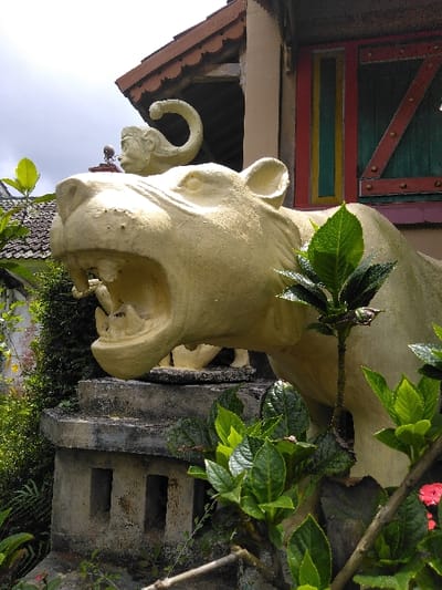 The image captures an outdoor scene in Ba Nwet Kone, Myanmar, featuring statuary and foliage against a building facade. A large, light-cream-colored concrete or stone statue of a roaring feline, resembling a tiger or lion with its mouth open and teeth visible, occupies the central foreground. This statue is positioned on a weathered, multi-tiered grey concrete pedestal with three visible rectangular openings. Behind the feline, a smaller, light-colored statue of an elephant's head with a raised trunk is partially visible. Lush green foliage, including plants with broad leaves and some small red flowers, surrounds the base of the statues and extends into the foreground. In the upper right background, a section of a building is present, distinguished by a reddish-brown scalloped tiled roof and a colorful window. The window features a red frame, a green panel with diagonal reddish-brown wooden slats on the right, and narrow vertical yellow and blue panels on the left. The scene is illuminated by bright, natural daylight under a partly overcast sky.