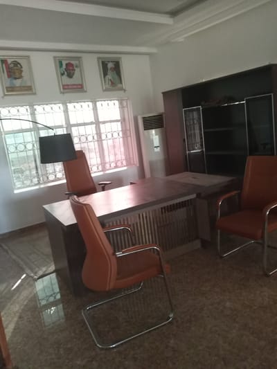 The image depicts an office interior, likely located in Hotoro North, Nigeria, based on provided context. Prominently featured is a large, dark wooden desk with ribbed detailing on its side panels. Two office chairs, upholstered in orange material with chrome legs, are positioned around the desk. One chair is placed in front of the desk, and another is to the right.
Behind the desk, a large, dark wood cabinet or bookshelf unit is visible, featuring glass-fronted doors and open shelving. To the left of the desk, a window with decorative metal bars allows diffused natural light to enter the room. Above the window, three framed portraits hang on the wall, each depicting a person in traditional attire. To the left of the window, a floor-standing air conditioning unit is present. The floor appears to be tiled with a speckled brown pattern, reflecting light from the window and casting shadows. The overall impression is of a somewhat formal office space.
