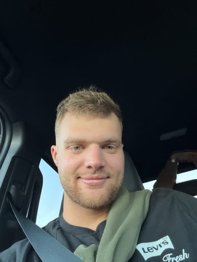 The image shows a Caucasian male with short blond hair and a light beard, seated in a car. He is wearing a black T-shirt with a Levi's logo and a green garment draped over his shoulder. A seatbelt is fastened across his chest. The car's interior is primarily black, with a glimpse of sky visible through the window. The image's location in Woodlawn, United States of America is unknown.