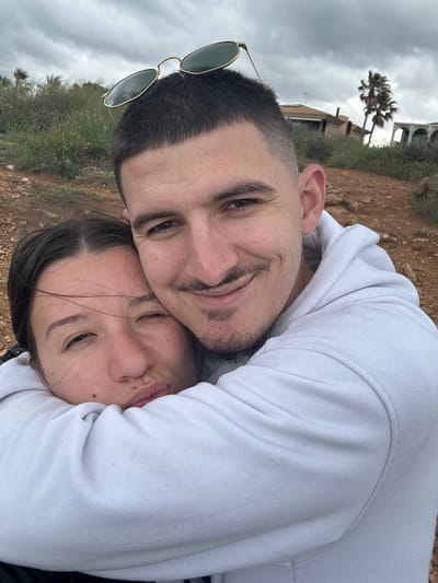 The image shows two people embracing in an outdoor setting, likely near a coastal area in Llucmajor, Spain. The man has short, dark hair and a beard, and is wearing sunglasses. The woman has dark hair. They are on a hillside with visible vegetation and a cloudy sky. In the background, buildings can be seen. The area is possibly a residential zone.