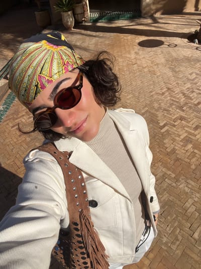 The image depicts a close-up, high-angle selfie of an individual located outdoors in Rissani, Morocco. The subject, appearing female, has short, dark curly hair and is wearing oval-shaped tortoise-shell sunglasses. Her head is covered by a multi-colored scarf featuring patterns in green, pink, yellow, and black. She is dressed in a light beige ribbed mock-neck top and an off-white denim jacket with visible dark buttons. A brown suede shoulder bag, adorned with silver stud detailing and fringe, hangs over her left shoulder. The subject looks directly at the camera with a subtle smile. The immediate environment consists of a reddish-brown brick or tile pavement laid in a herringbone pattern, strongly illuminated by direct sunlight, which casts distinct shadows. In the background, terracotta plant pots are visible, along with stone or mud-brick structures and a wall featuring a horizontal dark green tiled border.