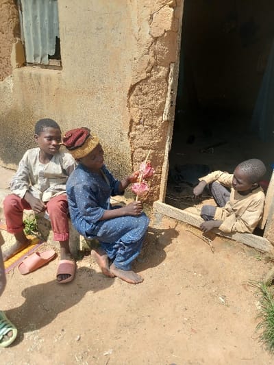 The image captures three children in an outdoor setting in Zangon Aya, Nigeria, positioned against a weathered building constructed of mud or earth. The structure features a window opening on the upper left covered by a corrugated metal sheet, and a dark doorway on the right side. The ground consists of dry, light brown earth, with sparse green vegetation visible in the lower right corner.
On the left, a child in a light-colored long-sleeved shirt and reddish-brown trousers sits on a low, concrete or mud ledge. Pink slide sandals are placed on the ground near their bare feet. This child observes the center. A second child, barefoot, wearing a dark blue patterned garment and a reddish-brown and yellow knit cap, squats on the ground in the center foreground. This child holds aloft a wooden stick with a multi-colored, pink and white confection. A third, younger child, dressed in a light brown long-sleeved garment, is partially visible sitting or crouching within the dark doorway on the right, looking towards the center child. A wooden plank forms the base of the doorway opening.