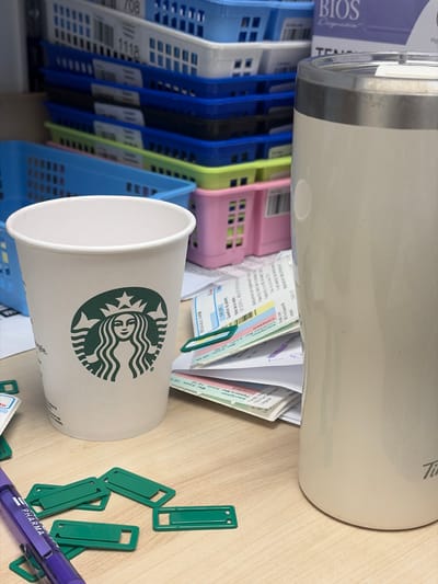 The image shows a close-up of a desk surface with several objects. In the foreground, a white Starbucks cup with the iconic green logo is prominently displayed on the left. To the right of the cup is a tall, cream-colored insulated tumbler. Scattered around these items are several green plastic clips and a purple pen with "PHARMA" written on it. Behind the cup and tumbler, stacks of blue, white, and pink plastic storage baskets are visible. Some of these baskets contain what appear to be documents or labels, with some text visible such as "708" and "1118". There are also some papers and receipts partially visible beneath the tumbler, one of which has text indicating dates and times such as "Mar 9 Juil, 00:00" and mentions "Longueuil". The overall environment appears to be an indoor workspace, likely an office or administrative area, potentially within the city of Longueuil, Canada, as indicated by the text on one of the documents.
