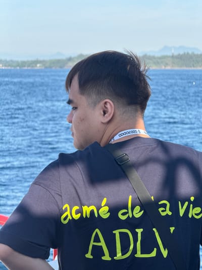 A medium shot captures a young man from the back, facing away from the camera, gazing out at a serene blue body of water. The man, with short, dark hair and a fair complexion, wears a dark navy t-shirt with bright yellow text. The text on his shirt reads "acmé de la vie" in cursive, followed by "ADLV" in larger, block lettering. A black strap of a bag is visible crossing his chest. Around his neck, a white lanyard with black lettering, partially obscured, is also visible. The background features a blurred landscape of lush green trees and distant hills under a clear, light blue sky. The water appears calm with gentle ripples, suggesting a tranquil environment, possibly a coastal area or a large lake. The overall scene conveys a sense of peaceful observation and contemplation by the young man.