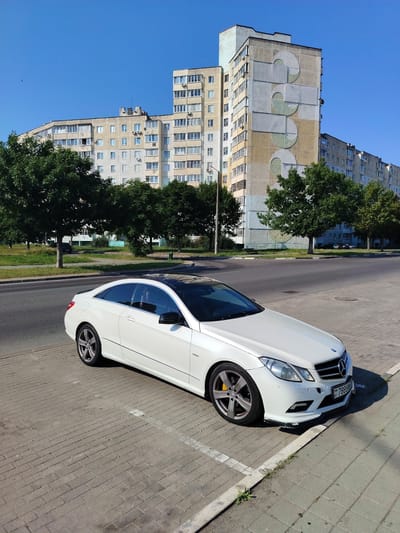 The image shows a white Mercedes-Benz E-Class coupe parked on a paved surface in front of a residential building. The car is positioned at an angle, with its front and right side visible. It has a black panoramic sunroof and grey alloy wheels. The license plate of the car is visible and reads "BY 7888 BK".
In the background, there is a multi-story residential building with a distinctly patterned facade, featuring large circular and rectangular shapes in yellow, white, and green. The building appears to be made of concrete. To the left of the building are several large, mature trees with dense green foliage. Beyond the trees, there is a clear blue sky with a single bird visible in the distance.
The scene is set on a paved area, possibly a parking lot or sidewalk, made of rectangular bricks. A road is visible to the left of the paved area, with a median strip and grass beyond that. The overall setting suggests an urban or suburban environment.
The lighting indicates it is daytime, with the sun casting shadows, particularly from the car. The weather appears to be clear and sunny. There are no people visible in the image. The overall mood is calm and static, with the focus on the parked car and the architecture of the building. The location context is Salihorsk, Belarus.