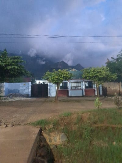 The image shows a view of a building and a street in Morogoro, Tanzania, located at coordinates -6.818, 37.696. The building is a single-story structure with a white facade and a dark green roof. It appears to be a residential home. The building has a gated front yard with a small tree in the front. There are electric power lines running overhead. The street is unpaved and is bordered by lush green vegetation. In the distance, there are mountains covered in clouds. The image appears to have been taken in the early morning or late evening as the sky is a deep blue. The scene is calm and serene, with no people or animals visible.