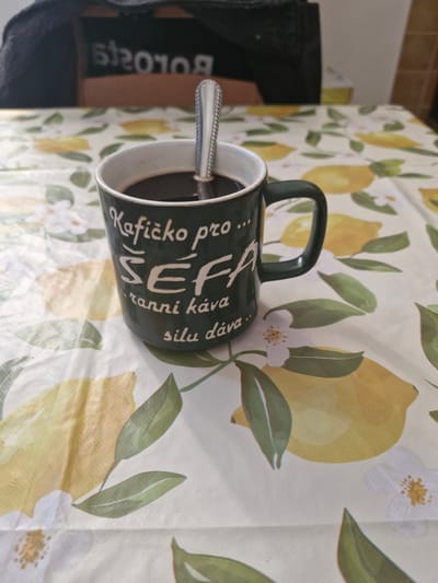 A close-up, eye-level shot captures a dark green ceramic mug filled with black coffee, with a silver spoon resting inside. The mug features white lettering that reads "Kafíčko pro... ŠÉFA ranní káva sílu dává." The mug sits on a white tablecloth adorned with a pattern of yellow lemons and green leaves. In the blurred background, a dark-colored garment is visible, possibly a jacket or sweater, draped over a chair. The lighting is moderate, casting soft shadows on the tablecloth. The scene appears to be set indoors, likely in a domestic environment. There is no indication of the specific location within Brno, Czechia.