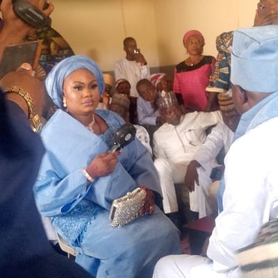 An indoor scene at Itakpe Junction, Nigeria, captures a gathering of individuals. In the foreground, a woman wearing a light blue traditional outfit, a matching headwrap, pearl-drop earrings, and makeup is seated, looking towards the camera. She holds a small black handheld fan in her right hand, which displays henna artistry, and a silver, beaded clutch bag in her left hand. Gold bracelets adorn her wrists. To her immediate right, the back of a man in a light blue cap and white attire is partially visible.

Behind the central woman, several people are present. A man in a white traditional garment and a patterned cap is seated, and a woman in a pink top with a black lower garment stands. Further back, numerous individuals, including several children, are seated. A young man in a white shirt stands further back, holding a dark object, possibly a phone or bottle, near his mouth.

On the extreme left, a person's hand wearing a gold watch holds a smartphone, whose back panel displays "Panasonic," indicating the device used to capture the image. The environment consists of plain, light-colored walls, suggesting a simple indoor setting.