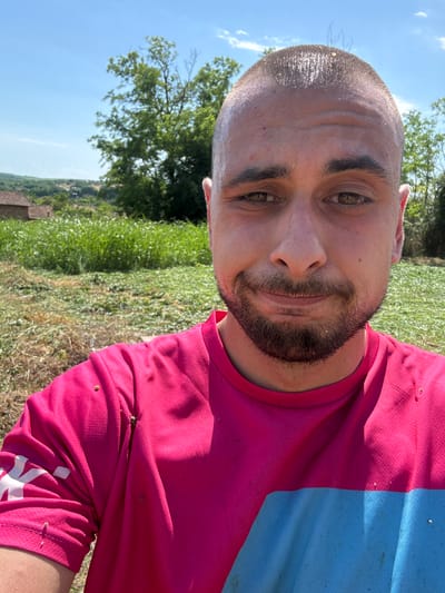 A man with a shaved head and a goatee poses for a selfie outdoors, possibly in Kozlovets, Bulgaria, given the rural landscape of rolling hills, greenery, and distant buildings. He is wearing a bright pink t-shirt with a blue stripe across the chest. The man's face is slightly sweaty, and there are some specks of dirt or debris on his head and shirt, suggesting he may have been engaged in outdoor labor or activity. The background is a bright, sunny day with a clear blue sky.