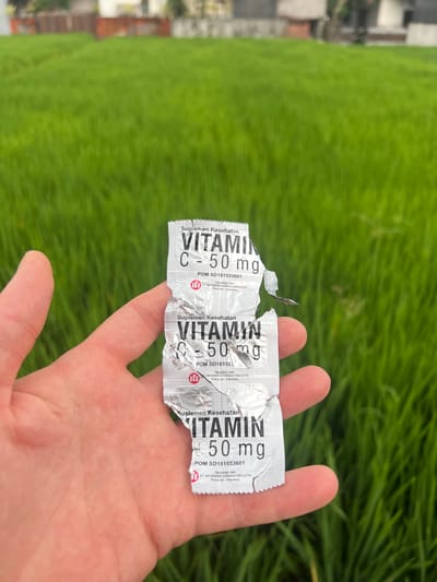 A hand holds a blister pack of Vitamin C supplements in front of a green, agricultural field. The blister pack has three doses, each labeled "VITAMIN C - 50 mg". The packaging is silver foil and partially torn open. The packaging also contains the words "Suplemen Kesehatan" and the registration number "POM SD181553601". Text on the packaging states that the product is manufactured by PT. IMFARMIND FARMASI INDUSTRI in Pasuruan, Indonesia. In the background, above the agricultural field, are buildings and rooftops, suggesting an urban or suburban fringe. The location is Celuk, Indonesia.