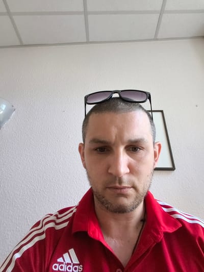 The image is a close-up, indoor selfie of a white male, appearing to be in his 30s or 40s. He has short, dark hair with some graying and a light beard and stubble. He is wearing a red collared shirt with white stripes on the shoulders and a white Adidas logo on the left chest. He has sunglasses perched on top of his head. Behind him, the wall has a textured finish and a white acoustic tile ceiling is visible. A framed picture with a dark border and a white mat is hanging on the wall to his right. To the left, a reflective, silver object, possibly a balloon, is partially visible. The lighting suggests it is daytime, likely from an overhead or natural light source. There is no identifiable information suggesting a specific location in Greifswald, Germany.
