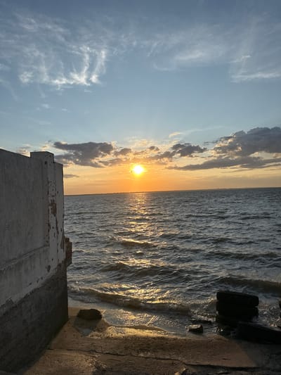 This image captures a sunset over the Black Sea in Zatoka, Ukraine. In the foreground to the left, a weathered concrete structure, likely part of a building or fortification, juts out into the water. Its rough texture and peeling paint suggest age and exposure to the elements. The lower section of the structure is a darker, more textured material, possibly stone or reinforced concrete.
The water itself is a deep blue-gray, with ripples and waves suggesting a gentle breeze. The setting sun, a bright orange orb, is positioned towards the center-right of the frame, casting a shimmering golden path across the water's surface. Above, the sky transitions from a clear pale blue near the horizon to a deeper blue overhead, dotted with scattered clouds. Some clouds are wispy and white, while others are darker and more substantial, tinged with orange and yellow from the sunset.
The shoreline in the foreground is a mixture of concrete and sand, with a large dark rock near the water's edge. To the far right, a stack of black tires rests partially in the water, a man-made element contrasting with the natural beauty of the scene. The horizon line is visible in the distance, with a faint, indistinct outline of land or buildings suggesting a settlement. There are no people or animals visible in the image.