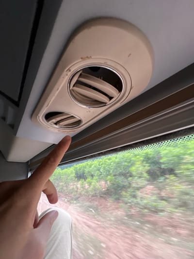 The image depicts the interior of a moving passenger vehicle, likely a bus or train, within or en route to Aê Thi, Vietnam. A light-skinned human hand, positioned in the lower-left quadrant, extends its index finger upwards, pointing towards a cream-colored overhead fixture. This fixture contains two circular air vents with visible adjustable slats, one larger than the other. The fixture exhibits visible wear, scuff marks, and dirt accumulation. Below the overhead fixture and the pointing hand, a window reveals a blurred exterior landscape, characterized by green foliage and reddish-brown soil, consistent with motion. Part of a person's leg or lap, covered in light-colored fabric, is visible in the lower-left.