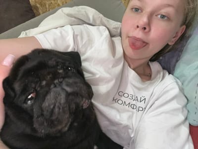 The image shows a young woman and a black pug lying down, seemingly on a bed. The woman is on the left side of the frame, her face turned towards the camera with her tongue sticking out playfully. She has light-colored hair and blue eyes. She is wearing a white t-shirt with text printed on it in black, Cyrillic letters. The text is oriented sideways and reads "СОЗДАИ КОМФОРТ". The pug is on the right side of the frame, its head resting on the woman's chest. The pug's face is mostly visible, showing its dark fur and eyes. The overall mood appears to be relaxed and affectionate, suggesting a casual moment between the woman and her pet. The lighting suggests it is daytime. The context mentions Klepiki, Russia.