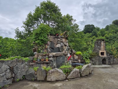 The image depicts an outdoor scene featuring a decorative rock structure resembling a waterfall, a stone barbecue grill, and a surrounding stone wall. Lush green foliage and trees form a dense backdrop. The sky is overcast with gray clouds. The rock waterfall is adorned with potted plants. The barbecue grill has a stone chimney and an opening with a brick interior. A concrete pathway or patio area is visible in the foreground. The overall impression is that of a garden or outdoor recreational area, possibly in a rural or suburban setting. The location is identified as Charnali, Georgia.
