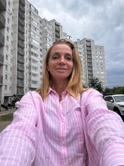 A woman with blonde hair, wearing a pink and white striped shirt, takes a selfie outdoors in front of a grey, multi-story apartment building. The sky is overcast and grey. A white car is parked to the right of the building. A person is walking away from the building on the left side of the frame. The location is the city of Kirishi, Russia.