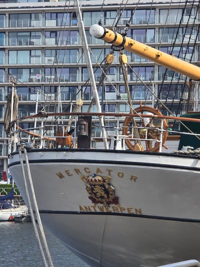 The image shows the bow of the Mercator ship in Ostend, Belgium, with a modern office building in the background. The name "Mercator Antwerpen" is displayed on the ship. A wooden steering wheel and a mast are visible. Ropes and rigging are interspersed with the geometric pattern of windows on the office building. A coat of arms is positioned beneath the ship's name. The ship is docked alongside the water, with a glimpse of nearby greenery.
