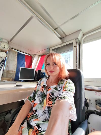 A woman with shoulder-length, light orange hair and fair skin is seated in a chair inside what appears to be a ship's bridge or control room. She is looking at the camera and smiling slightly, with a rosy hue on her cheeks. She is wearing a colorful, short-sleeved dress with a pattern of animal print, abstract shapes in green, orange, and black, and white accents.
The setting is a functional space with a white, paneled ceiling and white walls. To the left of the woman, on a desk, is a desktop computer monitor, keyboard, and mouse. Two circular clocks are mounted on the wall above the desk, with one displaying approximately 9:10 and the other an unreadable time. Behind the monitor, two flags are hung vertically, one red and one blue, with a white flag to the left. The window behind the woman offers a view of an outdoor scene with bright daylight, suggesting it is daytime. The weather appears to be sunny.
The chair the woman is sitting in is black and appears to be a captain's or operator's chair, likely for a vehicle like a boat or ship. To her right, part of the control panel is visible with some labels. One label clearly reads "220" in red. The overall impression is that the woman is on duty or taking a break while operating a vessel.