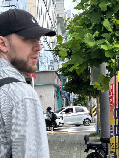 In Mokpo-si, South Korea, a close-up shot focuses on the left side of a man wearing a black baseball cap and a striped shirt. His face is partially visible, with a beard. Behind him, a light-colored hatchback car is stopped at a crosswalk. A woman with dark hair, wearing a hooded jacket and a backpack, stands near the car. In the background, there are buildings with glass windows and trees with green leaves. A lamppost with a sign is also visible on the right side of the image.