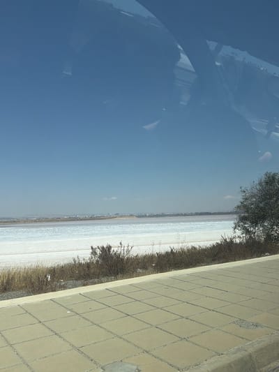 The image depicts a view of the Larnaca Salt Lake in Larnaca, Cyprus, as seen through a window. The foreground features a sidewalk paved with square tiles, some of which are cracked or damaged. Beyond the sidewalk, dry vegetation and brush grow along the edge of the salt lake. The salt lake itself appears as a vast expanse of white and light pinkish hues, suggesting a low water level and high salt concentration. In the distance, a city skyline with buildings is visible along the horizon, indicating the urban proximity of the salt lake. The sky is clear blue, with a few faint white clouds. Reflections of what appear to be parts of a vehicle's interior, such as a windshield frame and possibly seats, are visible on the window through which the photo was taken.
