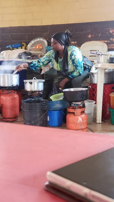 In Unguwar Shanu, Nigeria, a woman wearing a black headscarf and a blue and green patterned tunic is cooking outdoors. She is seated on a low stool, stirring a pot with a utensil. Several large metal pots and gas cylinders are arranged around her. Two red gas cylinders and one orange gas cylinder are visible, with pots placed on top of the cylinders to be heated. There are also several plastic buckets and basins nearby, some containing water or food items. In the background, a brick wall and wooden beams form the structure of the outdoor cooking area. The foreground is dominated by a red surface, possibly a table or a mat, with a smartphone resting on it. The scene suggests a communal cooking area or a busy street food vendor.