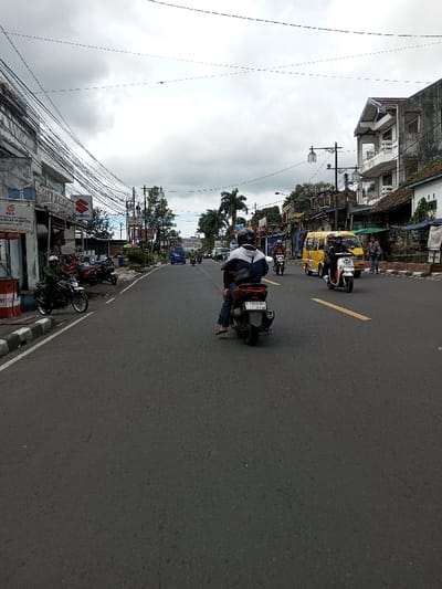 The image depicts an overcast daytime street scene in the city of Taohom, Laos. The perspective is from behind a dark-colored scooter, facing forward along an asphalt road.
In the immediate foreground, a person wearing a dark helmet, a blue and white hooded jacket, dark trousers, and light-colored sandals is seated on a dark scooter with the visible license plate "D 5157 NBY". The scooter is positioned slightly to the left of the road's center.
Along the road, traffic is moving in the same direction. To the right of the foreground scooter, a white scooter with two helmeted riders is traveling alongside a yellow minivan/microbus, which also contains at least two occupants. Further down the road on the right, additional vehicles and pedestrians are visible.
On the left side of the road, two motorcycles are parked near a curb next to a light-colored building. One black motorcycle has prominent chrome wheels. A person is seated on one of these parked motorcycles. Signs on the adjacent building include a "PKS" logo and a "SUZUKI" logo. A red and white barrel, potentially a fuel dispenser, is situated near the curb on the far left. Numerous dense overhead power lines and communication cables traverse the street, emanating from utility poles.
The right side of the street features various buildings of differing styles and conditions, including a prominent two-story building with balconies and a distinct roofline. Several streetlights are mounted on poles along this side of the road. Pedestrians are observed on the sidewalks.
The sky is heavily overcast with significant cloud cover. Palm trees and other vegetation are visible along the road in the mid-distance. The road surface is dark asphalt, marked with a solid white line on the left edge and a broken yellow line further to the right.