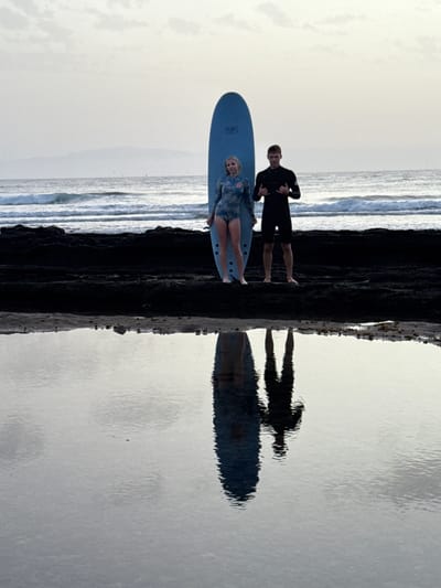 Two people, a woman and a man, stand on a dark, rocky shore with a surfboard standing upright between them. The woman, on the left, wears a long-sleeved swimsuit with a blue and white floral pattern. The man, on the right, wears a black wetsuit. They are both facing the camera. The ocean with small waves is visible behind them, and a distant, hazy coastline is in the background under a pale sky with scattered clouds. In the foreground, a calm pool of water reflects the figures and the surfboard. The scene appears to be on a beach near Arona, Spain, given the coastal landscape and the quality of light.