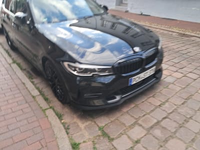 A black BMW sedan is parked on a cobblestone street in Parchim, Germany, with its front end angled towards the viewer. The car's license plate is partially visible, showing "PCH S370". The street is paved with reddish-brown cobblestones, and a narrow strip of grass runs alongside the curb. Buildings are reflected in the car's polished surface. The scene is captured from a slightly elevated, oblique angle, with motion blur suggesting the image was taken while the car was in motion or the camera was moving. The lighting indicates it is daytime.