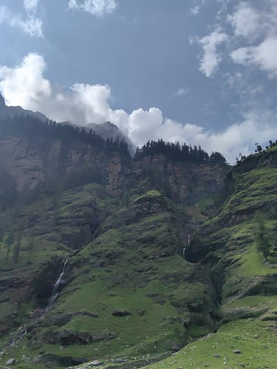 The image depicts a mountainous landscape near Kothi, India. The scene is dominated by steep, rugged slopes covered in lush green grass and dotted with scattered rocks. Several waterfalls cascade down the mountainsides, their white streams contrasting with the verdant terrain. A line of dark green pine trees forms a distinct band along the crest of the highest mountain peak, which is partially shrouded in clouds. The sky above is a pale blue, with scattered white clouds, some of which are dense and appear to be part of a mist or fog clinging to the upper reaches of the mountains. The overall impression is one of natural beauty and rugged wilderness.