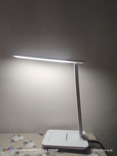 A white LED desk lamp is illuminated and angled towards a textured off-white wall, casting a bright light. The lamp has a rectangular base and a slender, upright arm supporting the light bar. The base has some subtle branding or control indicators, appearing as a series of lines. A black power cord extends from the right side of the base. The lamp sits on a surface covered with a patterned fabric featuring floral designs in muted colors. The image is watermarked with "Shot on realme 8 Pro 108MP" in the bottom left and the date "2026 06 23 14:45" in the bottom right. The scene appears to be indoors, likely in a domestic or office setting. The image does not contain any identifiable information about the city of Foligno, Italy.