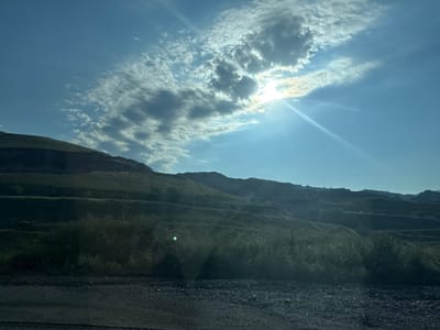The image depicts a scenic view of a sunlit sky with scattered clouds over a hilly terrain. The sun's rays pierce through the clouds, creating a bright, lens flare effect. The landscape appears to be a quarry or mine, with terraced slopes and sparse vegetation. The foreground is a dirt road or path, covered with gravel and small stones. The sky is a vibrant blue, with the sun casting a warm glow on the scene. The location is identified as the city of Arzni, Armenia. There are no people or specific objects of interest visible in the image, other than the natural elements of the landscape and sky.