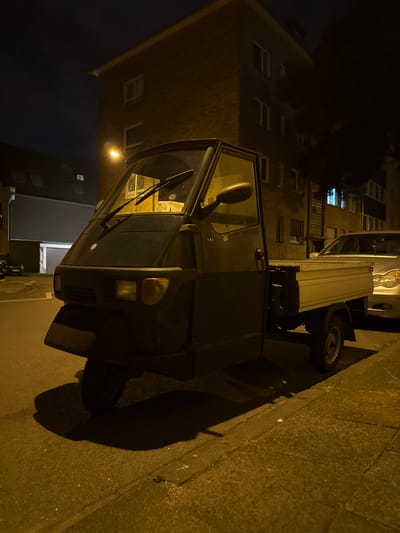 The image shows a three-wheeled vehicle, possibly an Ape, parked on the side of a street at night. The vehicle is dark colored and has an open cargo bed. The street is paved and there is a sidewalk. In the background, there is a building with several windows, and a street lamp is visible. The overall lighting suggests it is nighttime, with warm yellow tones dominating due to artificial light sources. The scene appears to be in a residential or commercial area within the city of Duisburg, Germany. The image is taken from a slightly low angle, focusing on the front and side of the vehicle. There are no people visible in the image.