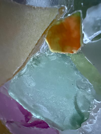 The image is a close-up, abstract view of colored glass fragments embedded in a clear, likely resin or epoxy material. The fragments are irregular in shape and appear to be of varying thickness and texture, some smooth and translucent, others rough and opaque. The colors present include a pale yellow, a vibrant orange, a soft green, and a deep pink. The overall impression is of a mosaic or a textured surface with distinct, colorful elements. The background is out of focus and appears to be a neutral gray. There are no people or discernible actions depicted in the image. The location in the city of Ridabu, Norway, is not evident from the visual information presented in the image.
