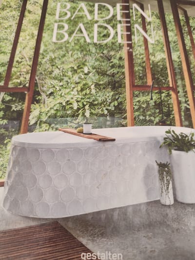 The image depicts a modern bathroom scene with a prominent bathtub. The bathtub is white and has a distinctive circular tile pattern around its base. A wooden tray is placed across the top of the tub, holding a white cup and a round object. There are two potted plants visible, one on the right side of the tub and another smaller one hanging near the floor. The background shows lush greenery, suggesting an outdoor or nature-inspired setting, framed by wooden beams. The words "BADEN BADEN" are visible in large white letters at the top of the image. The word "gestalten" is visible at the bottom right. There are no people in the image. The image is a photograph and does not appear to be located in Rosenheim, Germany.